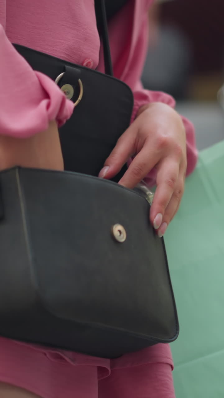 Young lady in pink dress and black top, riding an upward-moving escalator, searches through her black bag with concentration and a thoughtful expression, a mint green shopping bag rests by her side