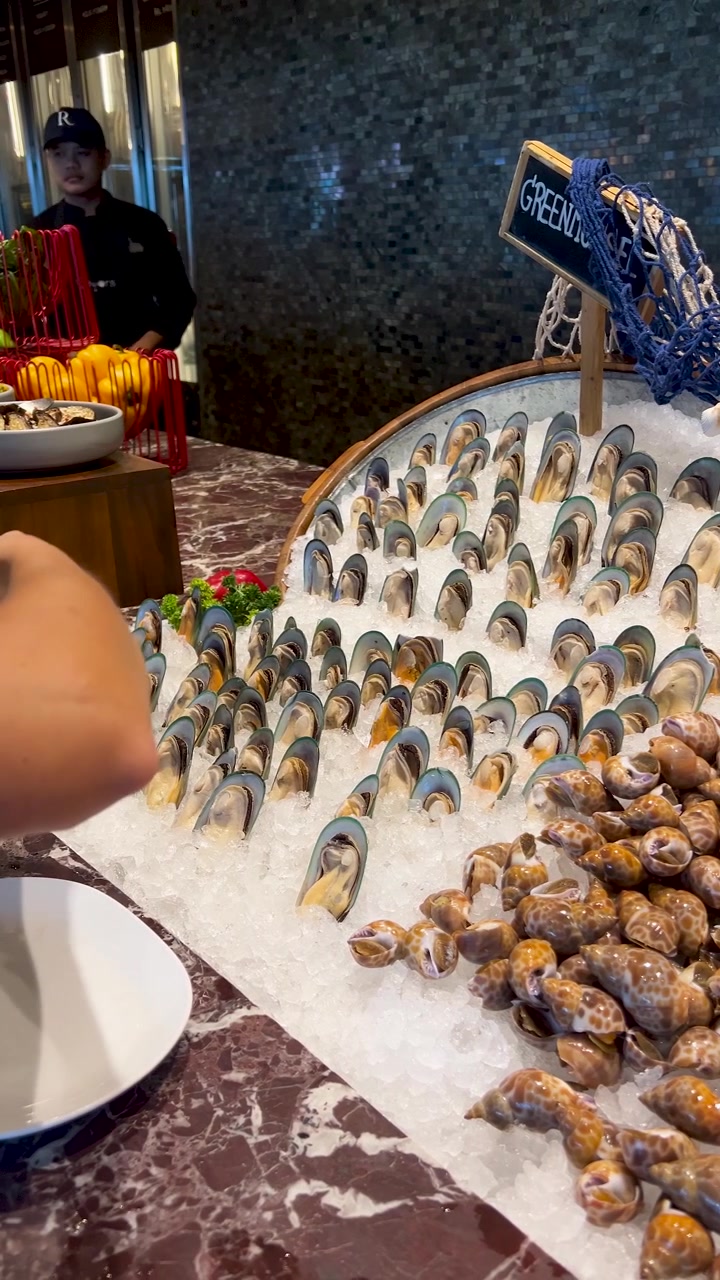 A person uses tongs to select fresh green mussels from a chilled seafood buffet display