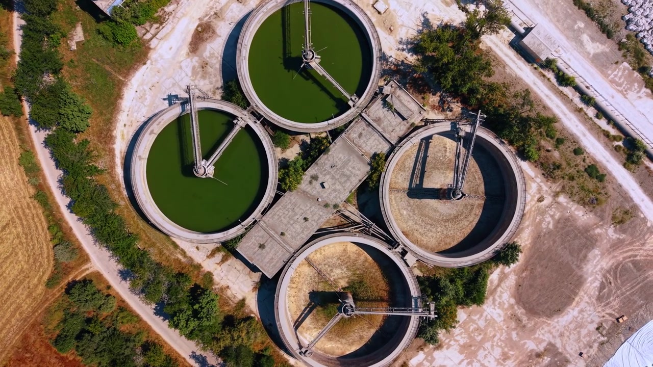 Top down view of circular industrial tanks. Two tanks contain green water while two dry basins reveal radial mechanical arms and concrete floors.