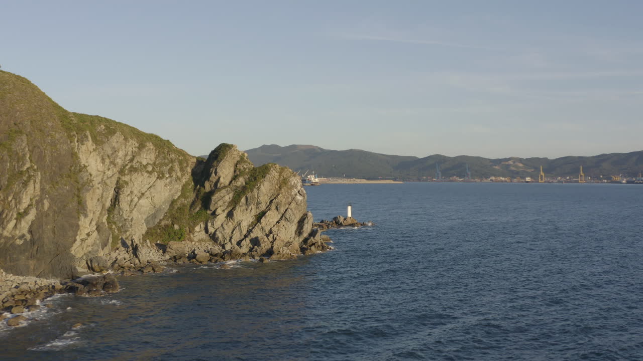 Inquadratura aerea rivelatrice di un terminal portuale per il carbone da dietro grandi scogliere a picco, con un piccolo faro bianco che si erge su scogli marini.