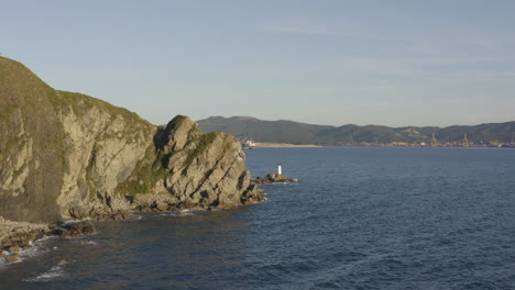 Aerial-reveal-shot-of-a-coal-port-terminal-from-behind-a-big-steep-cliffs,-with-a-small-white-color-lighthouse-beacon,-standing-on-sea-rocks