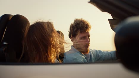 a bearded guy with curly hair in a blue shirt drives a white convertible together with a blonde girl and against the yellow sky