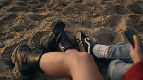 pareja de piernas sentadas en una playa de arena. chica de moda con un teléfono inteligente en la mano