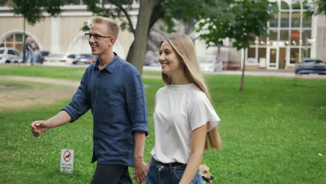 Una-Joven-Pareja-Positiva-Camina-Tomados-De-La-Mano,-Se-Encuentra-En-Un-Parque-De-La-Ciudad-De-Verano.-La-Niña-Y-El-Niño-Son-Rubios.-Feliz-Paseo-Por-El-Parque