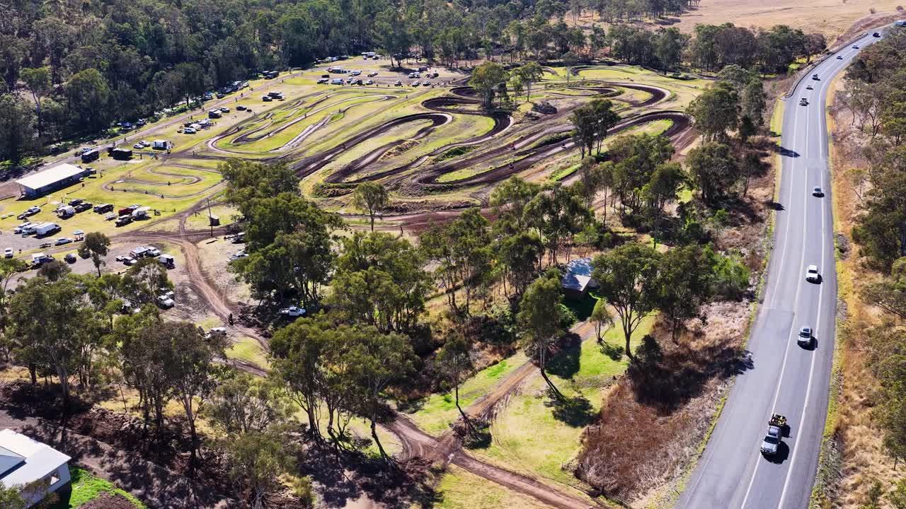 Drone footage pans over motocross park, cars driving on adjacent road, bright daylight, clear visibility