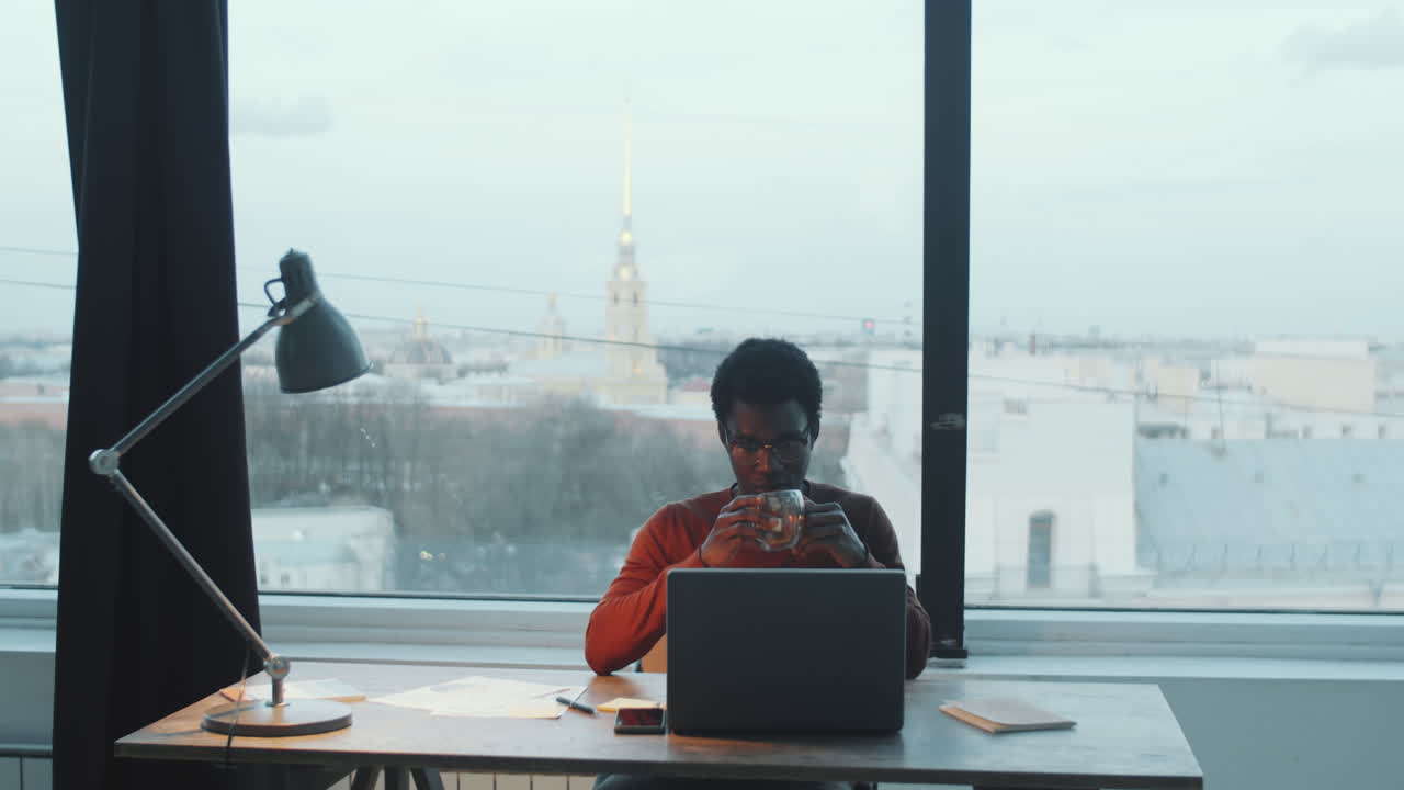 Uomo afroamericano che beve tè e lavora al portatile in un ufficio sul tetto