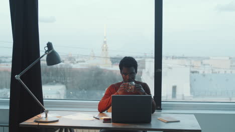 african american man drinking tea and working on laptop in rooftop office