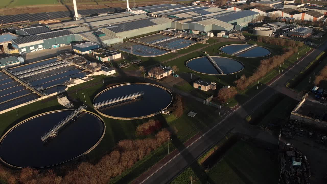 Backwards aerial view of a water treatment facility in The Netherlands showing the various circular and rectangular forms revealing the rotating blades of a wind turbine in the foreground