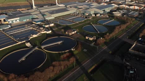 backwards aerial view of a water treatment facility in the netherlands showing the various circular and rectangular forms revealing the rotating blades of a wind turbine in the foreground