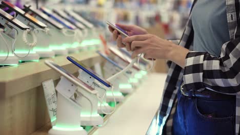 Waist-shot-of-unrecognizable-woman's-hands-chooses-a-smartphone-in-an-electronics-store.-She-takes-smart-phone-from-counter-and-trying-using-it.-Side-view