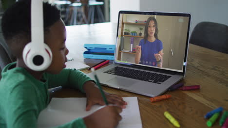 african american boy doing homework while having a video call with female teacher on laptop at home