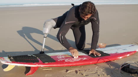 medium shot of a male surfer with prosthetic leg waxing surface of surfboard