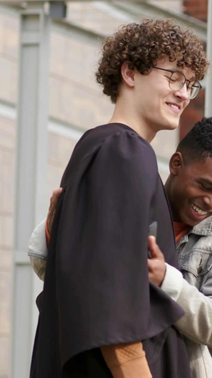 Vertical video: Taking cap from denim-jacket pal, grad in black gown fixing tassel outside building
