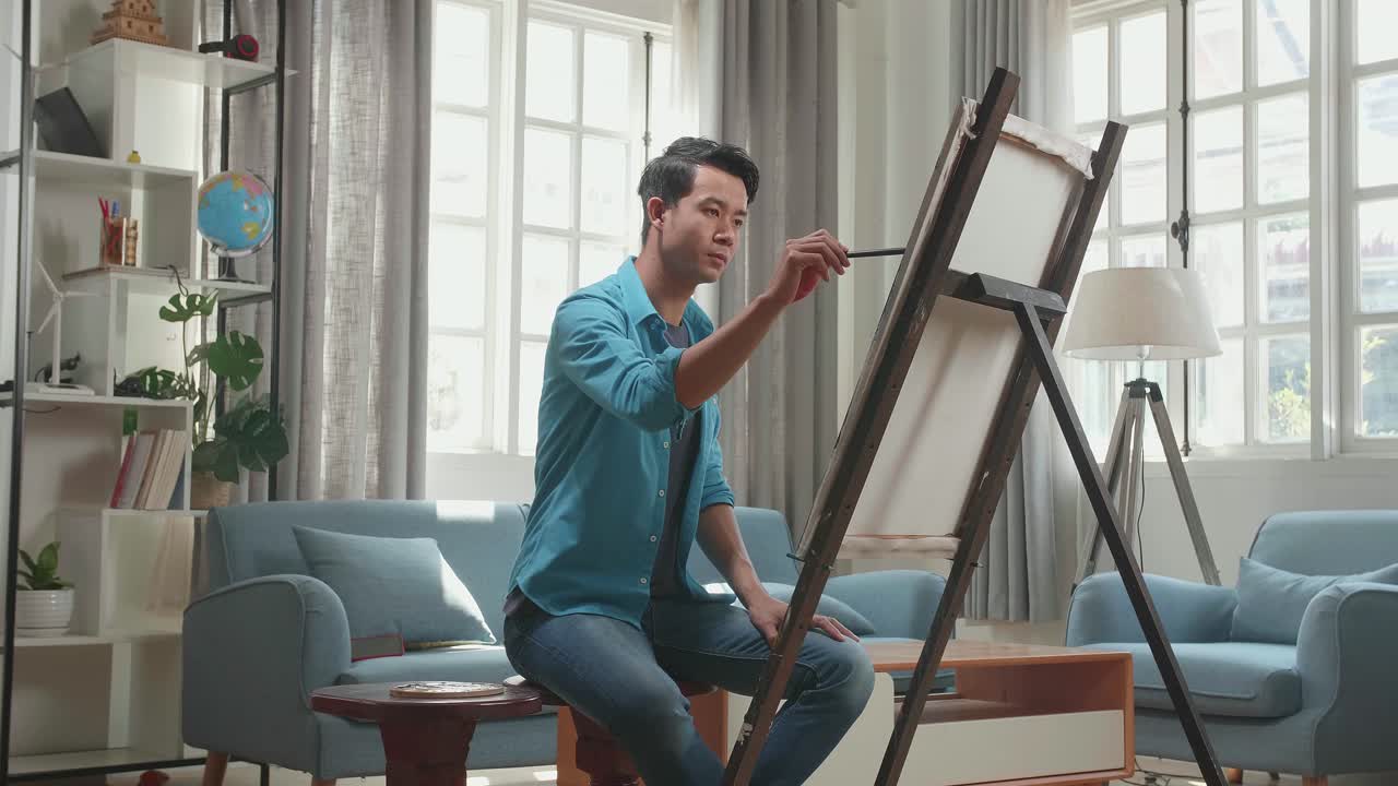 Side View Of An Asian Artist Man Sitting On Round Wooden Chair Without Backrest Holding Paintbrush Mixed Colour And Thinking Before Painting On The Canvas