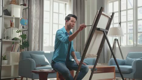 side view of an asian artist man sitting on round wooden chair without backrest holding paintbrush mixed colour and thinking before painting on the canvas