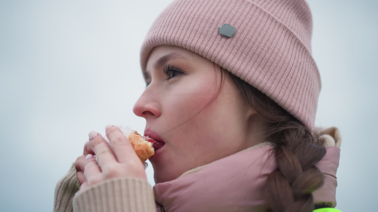 Close-up profile of young woman in pink beanie and scarf enjoying peaceful outdoor moment with eyes closed, holding sandwich, feeling calm and relaxed during winter day in cold weather environment