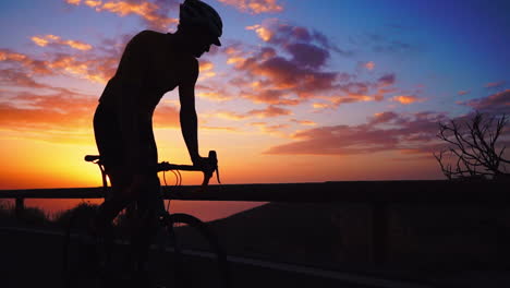 Silhouette-Slow-motion-reveals-the-athlete-cycling-on-a-mountain-serpentine,-appreciating-the-island's-view,-epitomizing-a-commitment-to-a-healthy-lifestyle-at-sunset