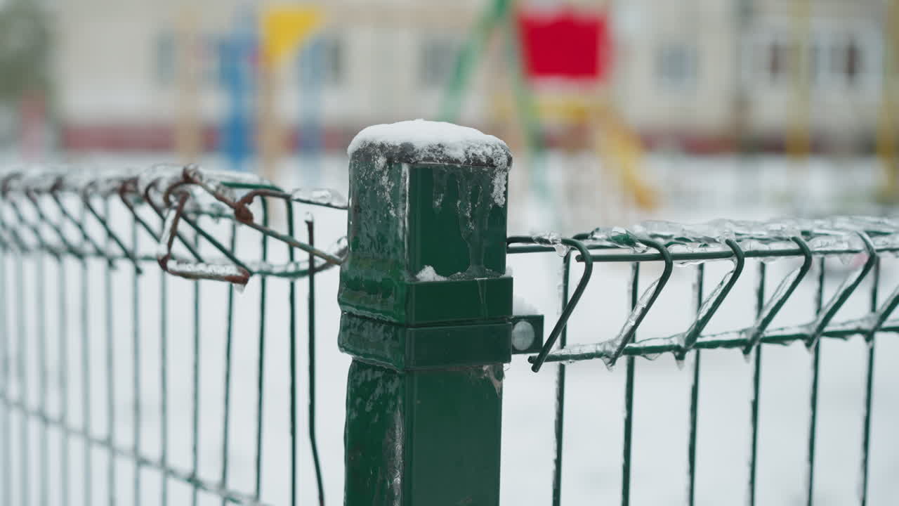 얼음으로 인 초록색 금속 울타리의 클로즈업, 와이어에 붙어있는 아이스클, 흐릿한 눈이 인 놀이터 배경과 멀리 있는 건물