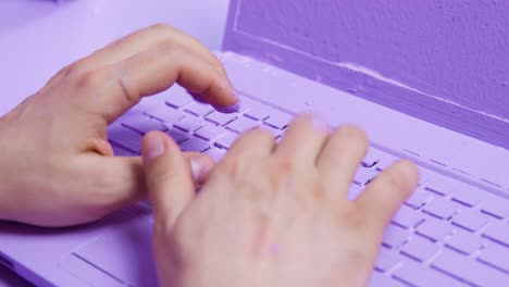 freelancer types text on laptop fully painted in purple color. hands of male person press buttons of device with painted keyboard and screen closeup