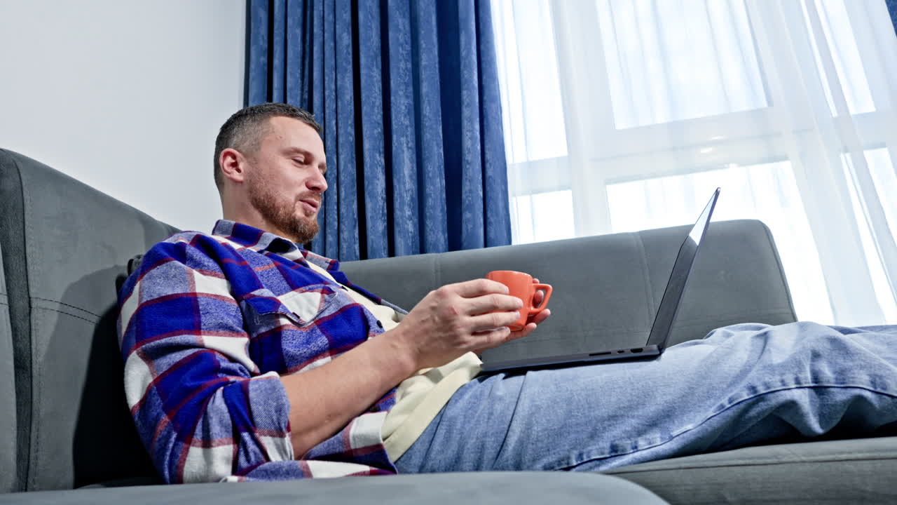 Man sits on couch using laptop while holding a cup in his hand during a sunny afternoon at home