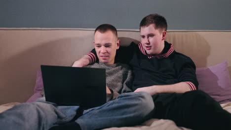 two men watching something on a laptop on a bed