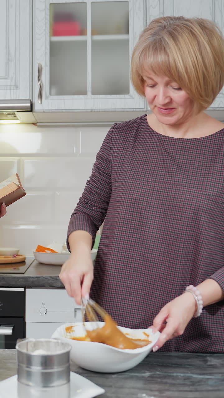 una joven lee atentamente un libro de cocina mientras observa a su madre agitando la masa en una cocina moderna, ella comprueba si la receta se sigue correctamente