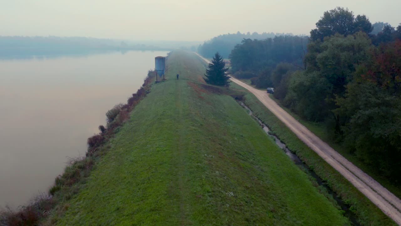 vista aérea de un hombre parado junto a un silo de metal en una colina cubierta de hierba con vistas a un lago brumoso y reflectante