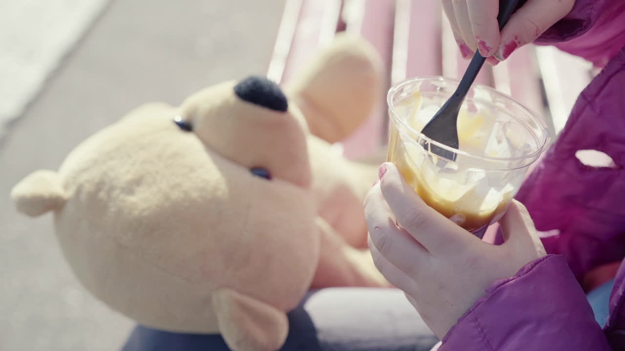 파란색 점퍼 매니큐어와 테디베어를 들고 야외에 앉아 있는 선디 아이스크림을 들고 있는 어린 소녀의 클로즈업