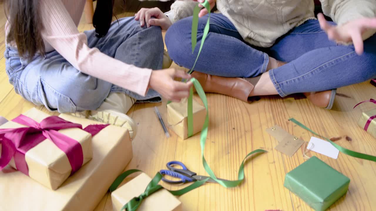 Madre che prende un nastro verde e guida la figlia a fare un fiocco su una scatola regalo a casa, pronta per essere donata.