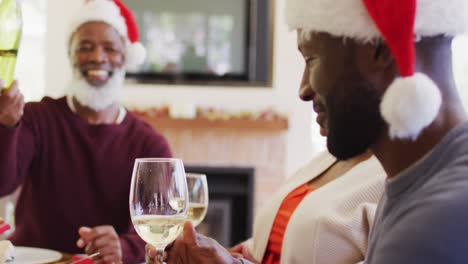 Afroamerikanischer-älterer-Mann-Mit-Weihnachtsmütze-Gießt-Getränk-In-Ein-Glas-Seines-Sohnes,-Während-Er-Auf-Dem-Esstisch-Sitzt