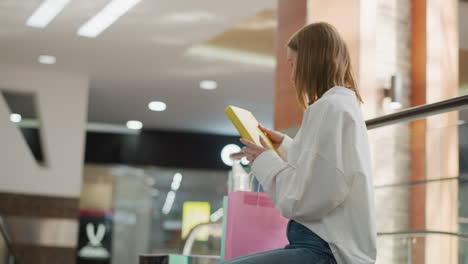 side view of a woman seated in a shopping mall, retrieving a yellow-covered book from a pink shopping bag, surrounded by a vibrant modern interior with glass railings and a bright atmosphere