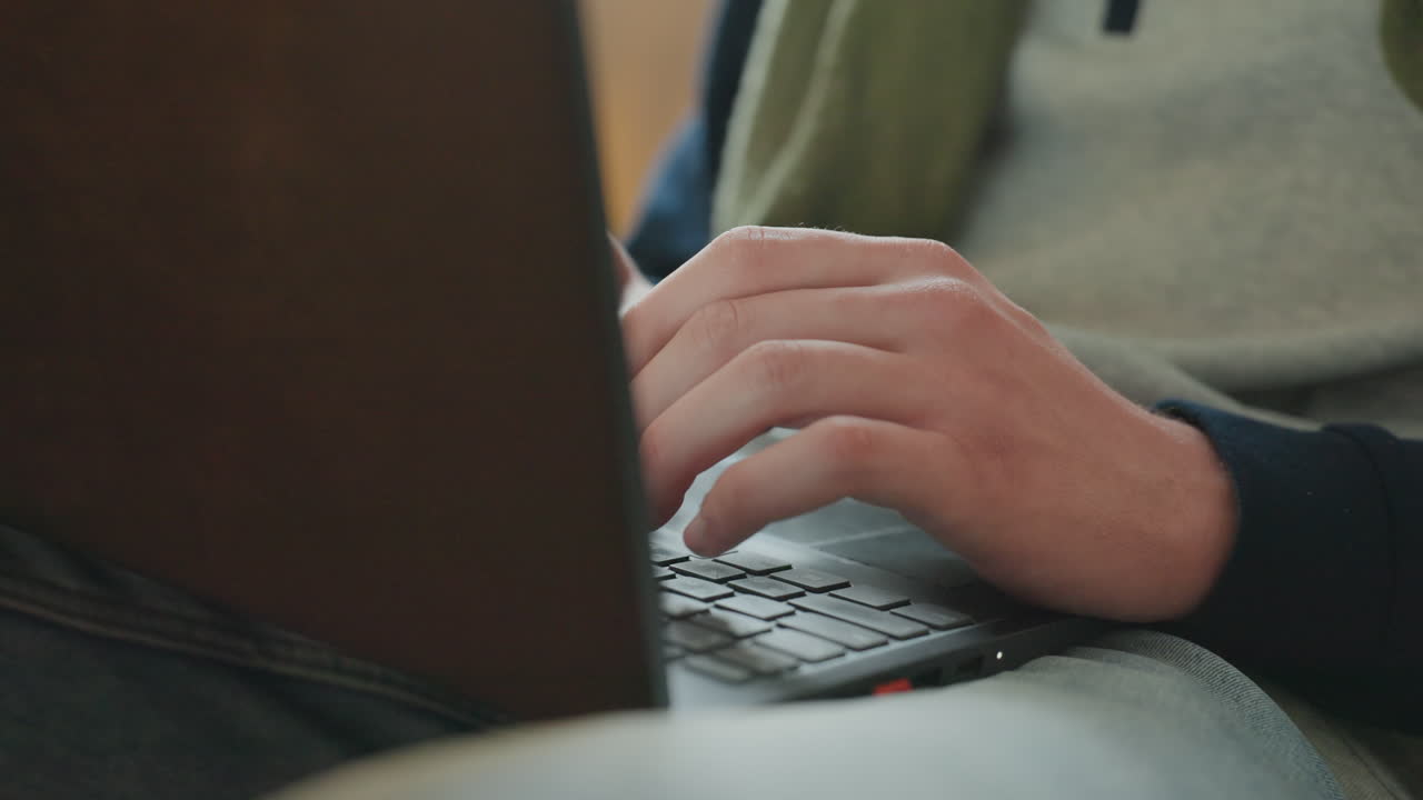Nærbillede af hænder, der taster på bærbar på sofa, hyggelig indendørsscene med grøn sweater og denimjeans, varme bokeh-lys i baggrunden, fokuserede fingre trykker på taster, freelance skribent og studerende arbejder.
