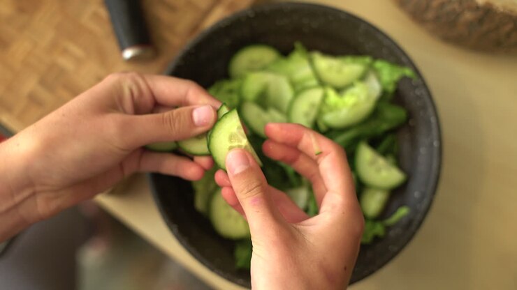 Close Up of Female Hands Adding Sliced Cucumber to Bowl of Lettuce