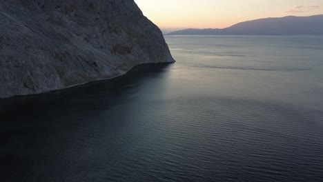 Steep-rocky-cliff-leading-into-ocean-water-before-sunlight-in-the-background,-aerial-footage,-dolly-out,-reveal