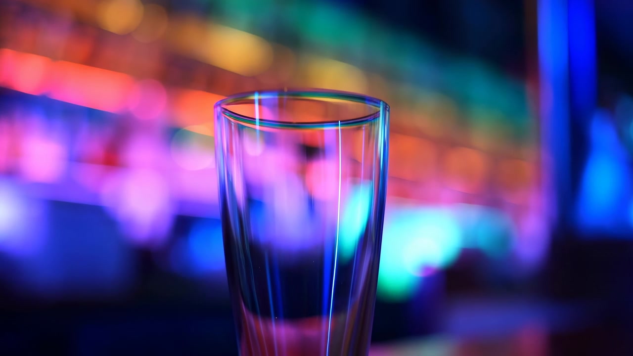 Primer plano de un solo vaso de chupito vacío centrado en una encimera de bar brillante, enmarcado contra estantes borrosos retroiluminados con neón y botellas desenfocadas. Poca profundidad de campo.