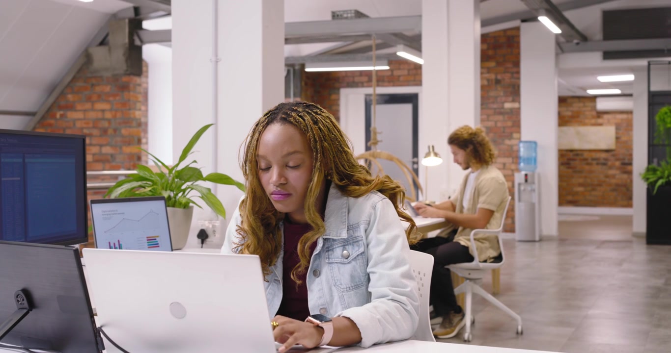 Adult African American woman entering office holding cup placing drink on coaster, typing on laptop