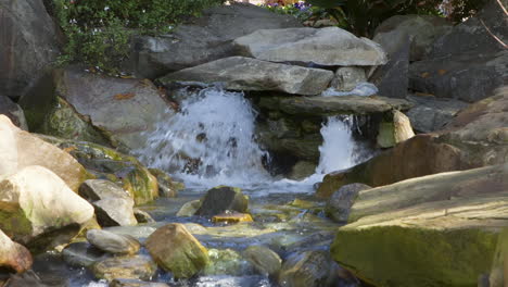 Small-waterfall-flowing-into-fresh-water-stream-and-rocks-of-various-sizes-and-shapes,-during-a-clear-and-sunny-afternoon