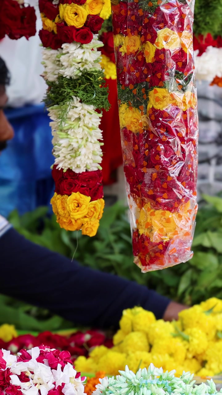 La mano di una persona si allunga verso una vivace ghirlanda di fiori di calendula tra colorate composizioni floreali in un mercato all'aperto a Little India, Singapore. Luce naturale, vista ravvicinata