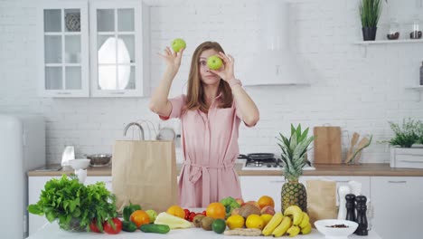 funny young woman closes her eyes with two green apples indoors. beautiful lady holds apples on her face and looks at camera in the light kitchen. healthy food concept.