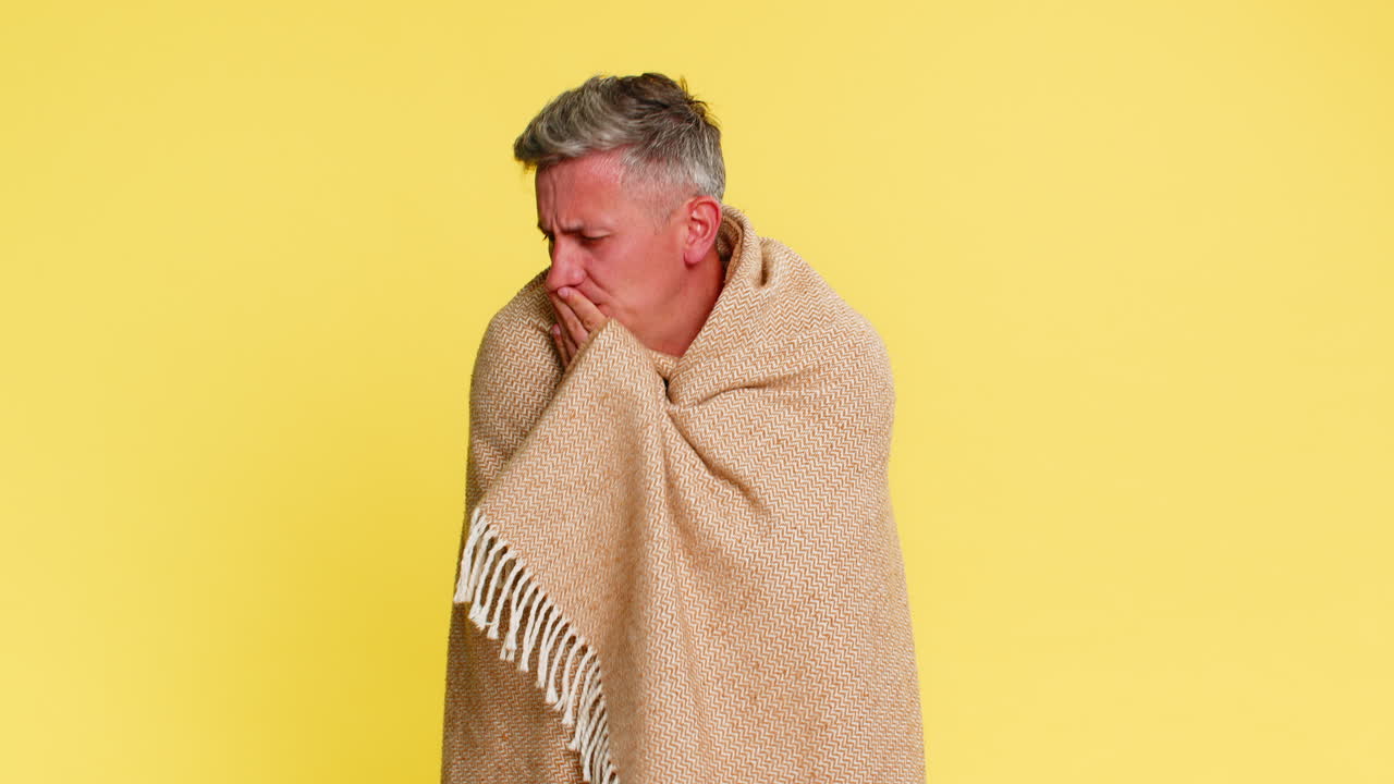 Adult man wrapped in blanket coughing with tired sick face showing flu symptoms cold virus illness
