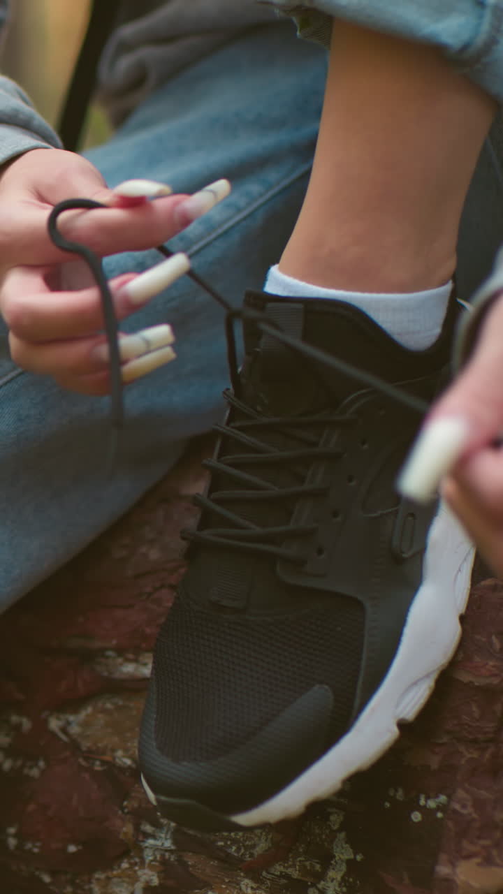 Close-up of woman tying shoelaces on black sneakers, wearing gray sweater and light blue jeans, with long white nails, seated on fallen tree in forest