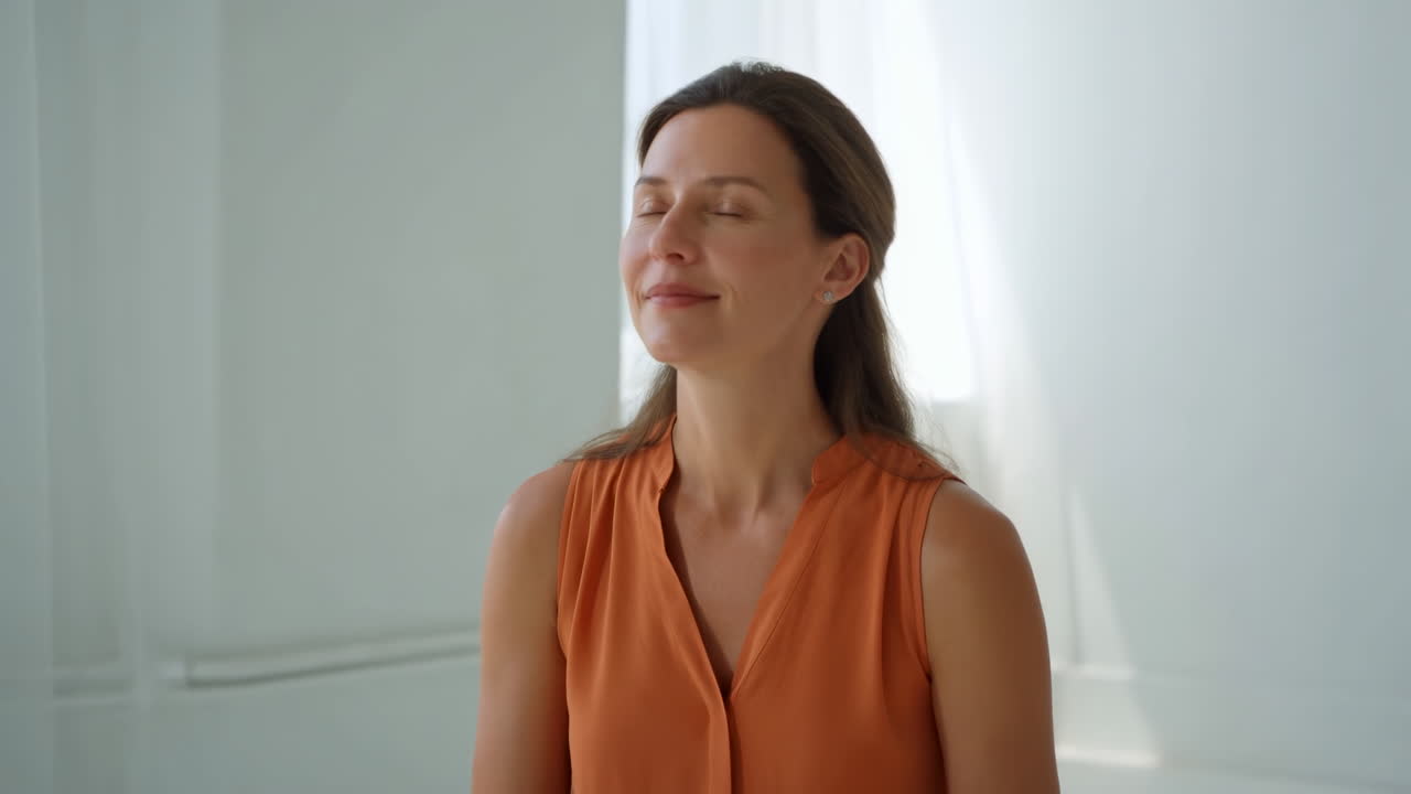 Donna bianca in studio illuminato dal sole che pratica la mindfulness, occhi chiusi, sorriso delicato, esercizio di respirazione lenta, arancio.