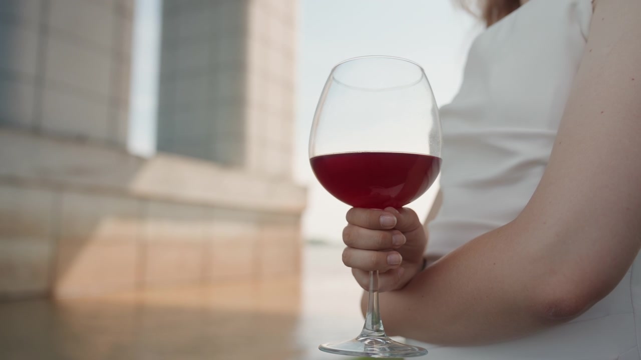 Una donna in abito bianco tiene e muove leggermente la mano con un bicchiere di vino contenente liquido rosso. Lo sfondo presenta una struttura architettonica che suggerisce un ambiente esterno o semi-esterno con luce soffusa.