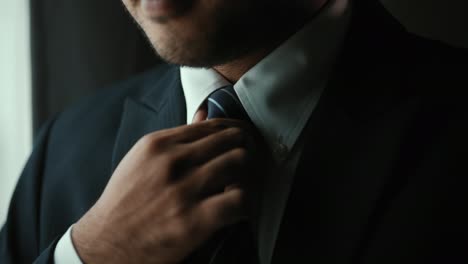 confident businessman adjust classic blue suit with necktie near window in hotel room. handsome man wearing a nice suit on wedding day.