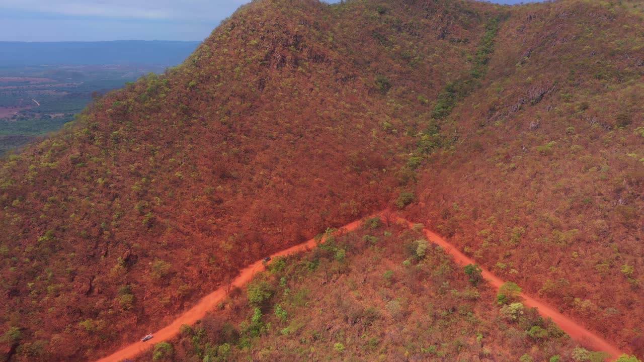 High drone shot follows lone gray 4x4 crew cab pickup truck threading along a red dirt road in the valley between two steep Cerrado slopes on the route to Tocantins, Brazil