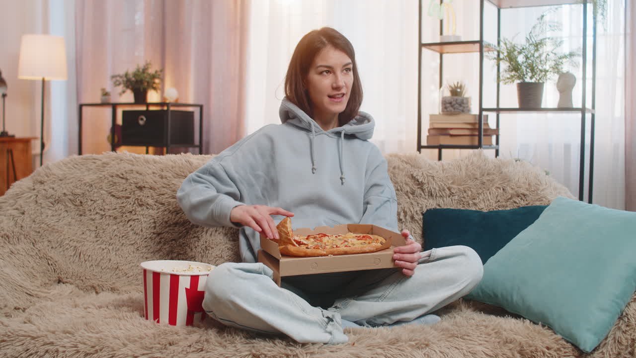 Giovane donna che mangia una deliziosa pizza e popcorn (cibo spazzatura) seduta sul divano a guardare la TV a casa