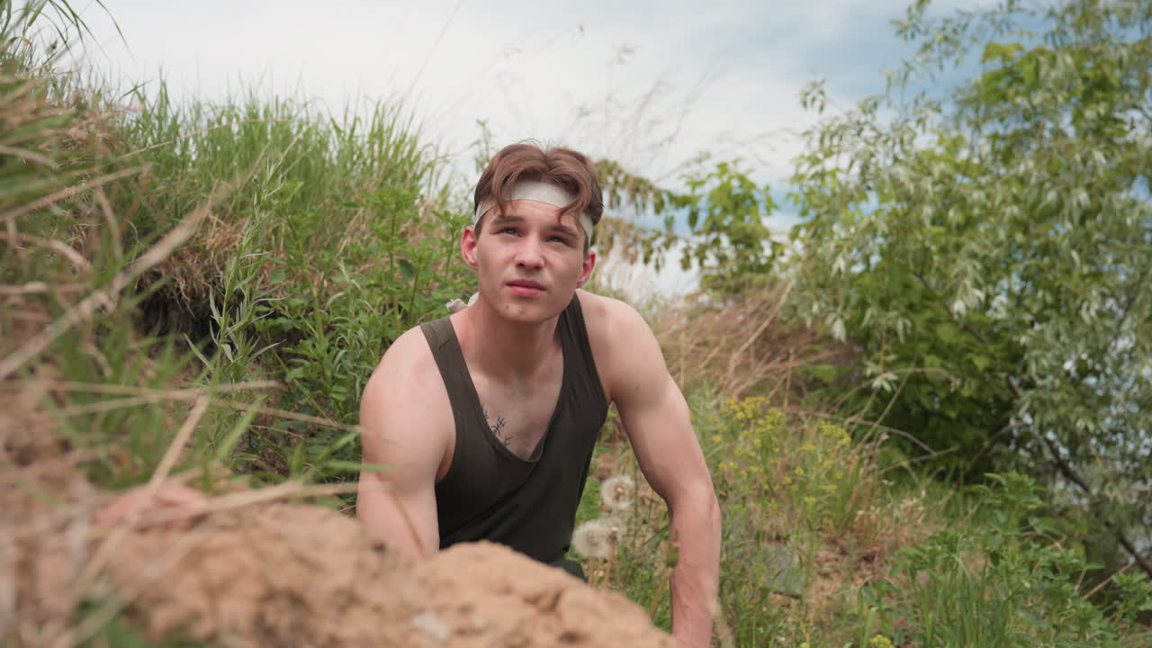 Adventurer sitting on steep grassy slope near river shaking head while gazing at cloudy sky catching breath after strenuous climb in serene wilderness setting with tall grasses and bushes