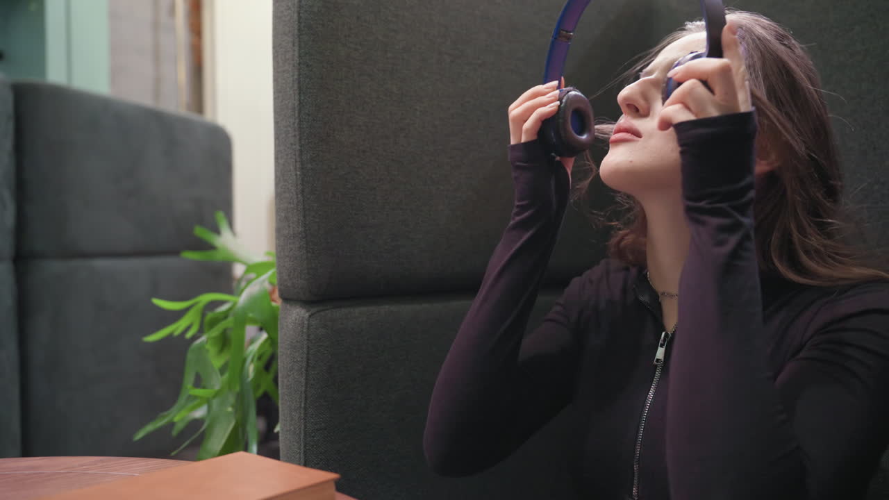 Light skin lady with long brown hair sits beside cushioned wall, gently closes brown book and begins removing headset while seated at wooden table in calm indoor setting with green plant in background
