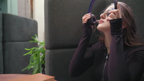 light skin lady with long brown hair sits beside cushioned wall, gently closes brown book and begins removing headset while seated at wooden table in calm indoor setting with green plant in background