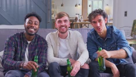 multi-cultural group of male friends on sofa at home drinking beer and doing cheers to camera